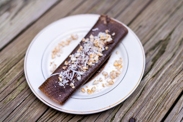 Closeup of chocolate vegan banana crepe dessert wrap with rice crisps, coconut flakes made in dehydrator on plate wooden table