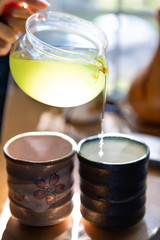 Closeup of person hands holding glass teapot pouring Japanese sencha green tea in cups or teacups at home for breakfast morning by window