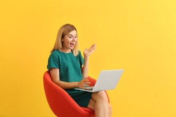 Shocked woman with laptop sitting in armchair on color background