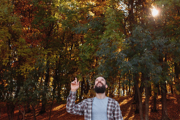 Fall forest. Happy hipster guy pointing finger up in air in nature park. Autumn trees background.