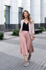 a young girl walks along the sidewalk in wide strides. Skirt plise, long jacket, sneakers. Long wavy hair. Very beautiful European in the summer walks down the street in a great mood.