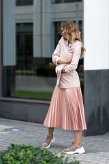 girl model in coral color clothes. Pleated long skirt, jacket, sneakers, black blouse. Long wavy hair. A very beautiful European woman stands on the street near a glass case.