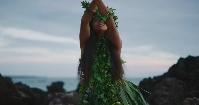 Traditional Hawaiian hula dancing at sunset in slow motion, woman performing Hawaiian hula with haku leis and ti leaf skirt with the ocean in the background