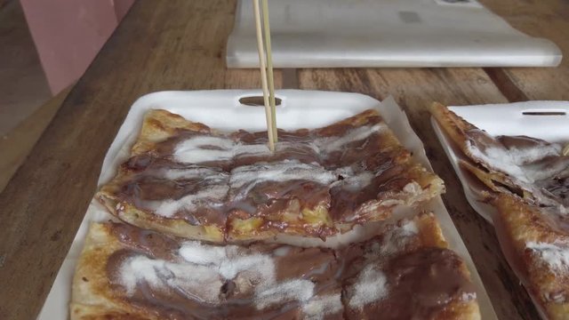 Extreme close up of wood stick pick a slice of delicious freid pancake whit chocolate, coco and condensed milk. Tyical Lao street food in take away in a plastic box. Vang Vieng, Laos.