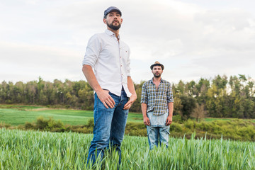 Two men in plaid shirt standing in oat field with forest in background posing