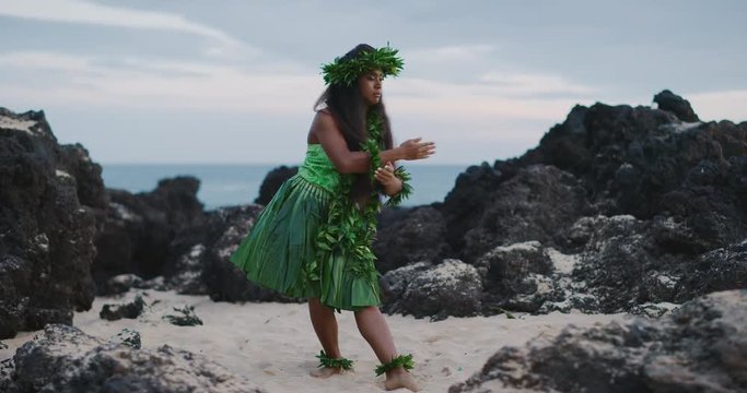 Traditional Hawaiian hula dancing at sunset in slow motion, woman performing Hawaiian hula with haku leis and ti leaf skirt with the ocean in the background