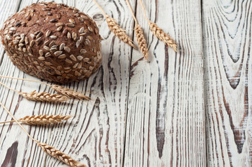 One whole loaf of rye bread with seeds of sunflower near spikelets of wheat lies on old weathered white wooden table