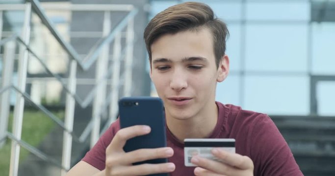 Teen makes online purchases using a credit card and mobile phone.  Smiling young man makes online shopping using cell phone with bank card, outdoors. Boy uses smartphone and credit card. Real time.