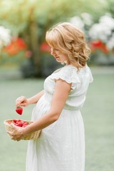 Young beautiful pregnant woman in a white dress walks through the park and eats strawberries. Picnic. Motherhood.