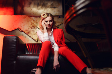 Full-lenght photo of young blonde in red jacket sitting on leather sofa