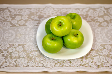 Green apples served on white porcelain plate on a table covered with white lace towel.