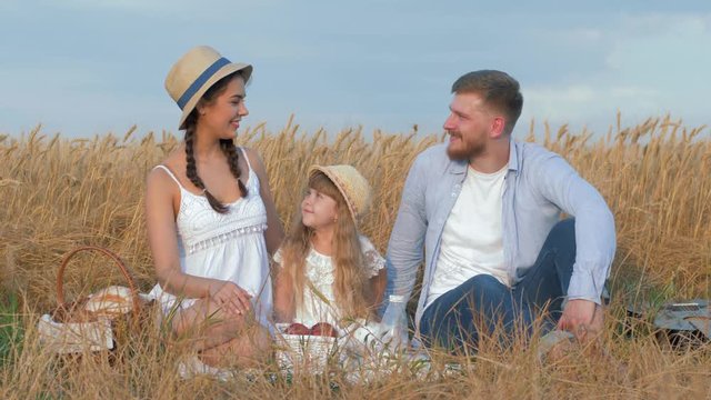 portrait of young family outdoors, happy couple sits with their little daughter smiling at open air picnic in autumn golden grain wheat harvest seasonal field