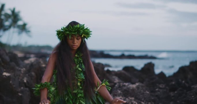 Traditional Hawaiian hula dancing at sunset in slow motion, woman performing Hawaiian hula with haku leis and ti leaf skirt with the ocean in the background