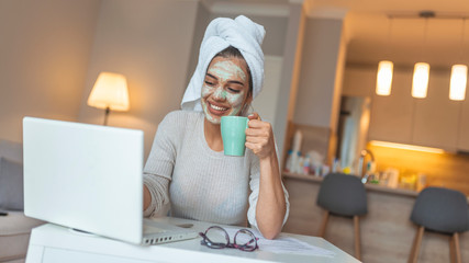 Portrait of beautiful woman with facial mask taking a coffee at home while use internet with modern laptop computer - Home scene for modern lifestyle people.