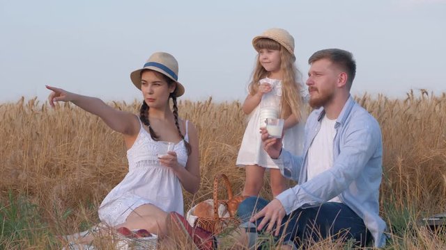 weekend family picnic, happy young mother looks and points towards side talking to husband and little daughter during outings with baked buns and milk in grain wheat meadow at harvesting against