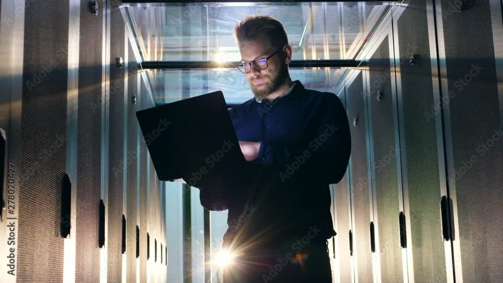 Poster it support engineer in glasses is operating a laptop in a server room