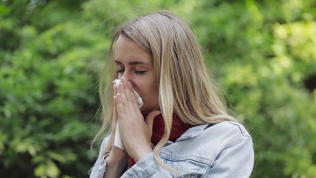 Young woman with with allergy symptom blowing nose standing in the park. Sick girl sneezing and blowing her nose into tissue due to allergy to tree pollen.