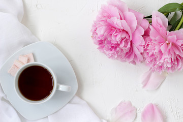 Stylish breakfast, a cup of tea, a tablecloth, flowers peonies and petals on a white table. Morning mood. bright serving meals, breakfast alone in the bright sunshine.