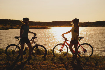 Two silhouettes of women with bike standing on lakeside and talking each other at the sunset