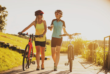 Two female friends walking with bikes and talking each other against the sunset light