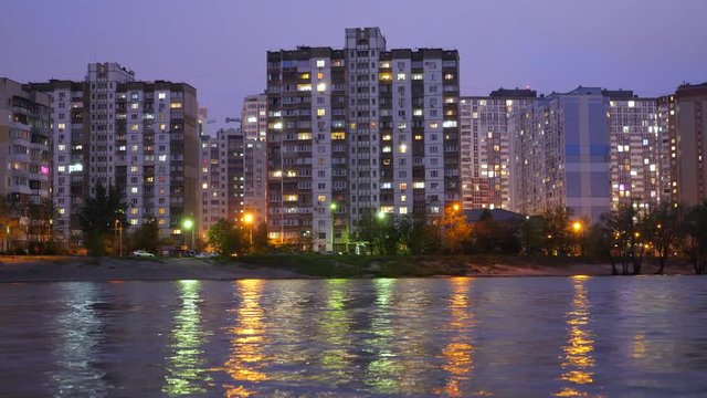 Appartment buildings located near the lake, during sunset with purple sky reflecting in the water. Electricity consumption. Time lapse. Evening city lights reflecting in the water, river, lake.