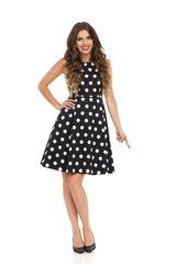 Beautiful Young Woman In Black Cocktail Dress In Polka Dots And High Heels Is Pointing Down And Smiling