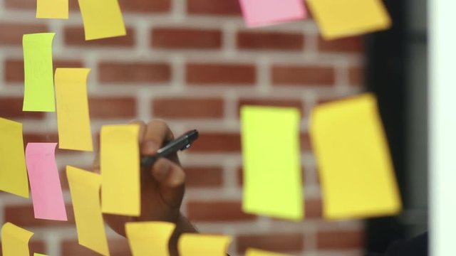 Asian creative man stick a sticky note on glass board. Young professional business male write information, strategy, reminder on paper, business situation, startup in Loft office concept. Slow motion.
