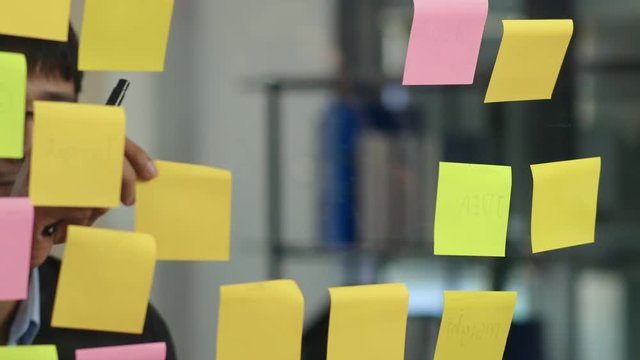 Asian creative man stick a sticky note on glass board. Young professional business male write information, strategy, reminder on paper, business situation, startup in Loft office concept. Slow motion.