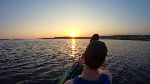 Right behind shot: Man paddling a kayak at sea moving towards the setting sun