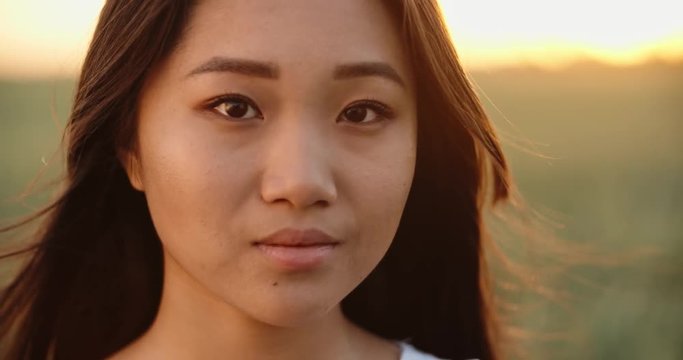 Portrait shot of panasian girl standing in golden wheat field during bright sunset, with wind blowing ito her hair - close up 4k footage