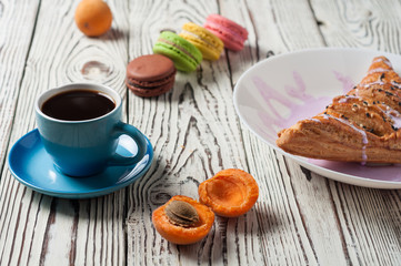 Fototapeta na wymiar Full ceramic cup of hot black coffee near baked sweet bun in form of triangle with purple cream, apricot, macaroons lies on white weathered wooden table