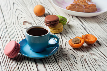 Full ceramic cup of hot black coffee near baked sweet bun in form of triangle with purple cream, apricot, macaroons lies on white weathered wooden table