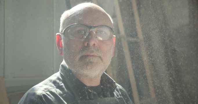 Close-up portrait of senior male carpentry master working at wood factory watching into camera being serious and calm.