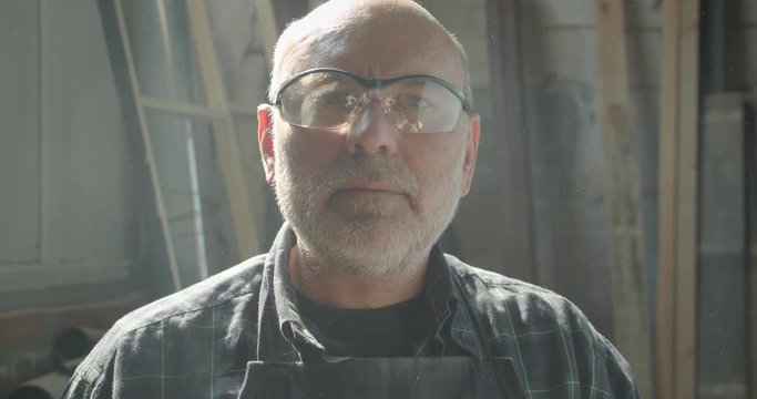 Close-up portrait of senior carpentry master in safety glasses working at wood factory watches into camera being serious.