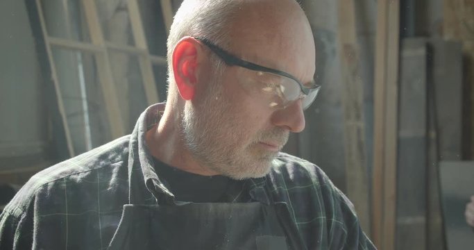 Close-up portrait of senior carpenter in safety glasses controlling the process at wood factory watches into camera being calm.