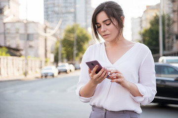 Beautiful brunette business woman in white skirt and grey suit trousers working on a mobile phone in her hands outdoors. European city on background. copy space