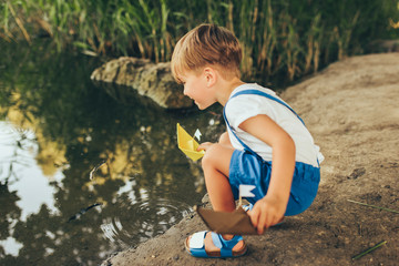 Image of cute little boy launch paper ship on the lake in the park. Adorable kid boy playing with a boat. Curious child sailing a toy boat by the waters' edge outdoors. Childhood and ecology concept