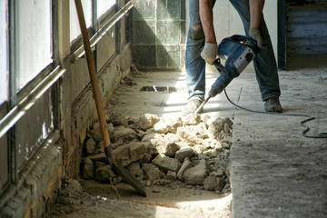 A man using a perforator dismantles the cement floor in a room with large windows