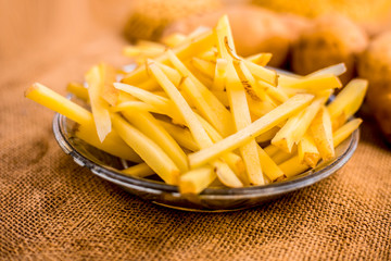 Raw cut french fries in a transparent glass plate along with raw potato with it on jute bag's surface.