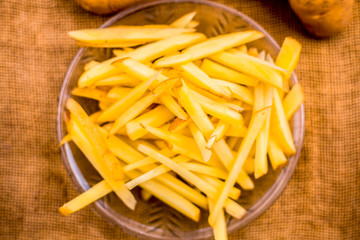 Raw cut french fries in a transparent glass plate along with raw potato with it on jute bag's surface.