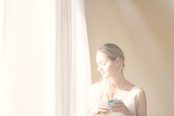 Young beautiful women drinking coffee in the mornng bedroom near window with white curtain overlay. Happy caucacian girl having a good time drinking tea.