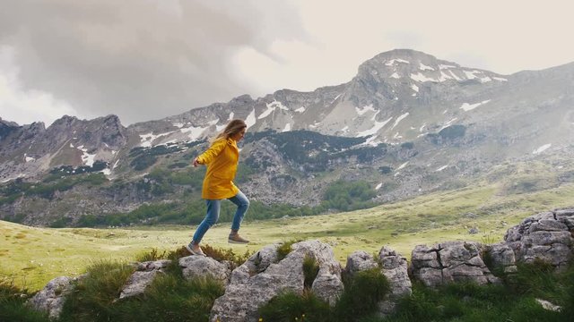 Young cheerful woman in a yellow raincoat running and having some fun on rocks with beautiful mountains background, slow motion