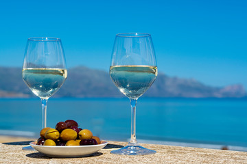 Two wine glasses with white wine served with olives on outdoor terrace witn blue sea and mountains...