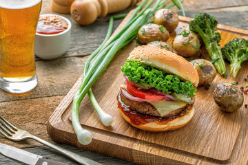 Hamburger with grilled vegetables and scallion served on a wooden board and a glass of beer on a rustic tale.