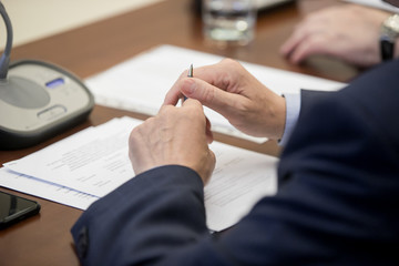 A politician or businessman in a suit sits at a polished wooden table in front of a microphone...