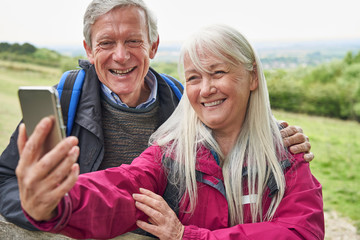 Smiling Senior Couple Hiking In Countryside Standing By Gate  And Taking Selfie On Mobile Phone
