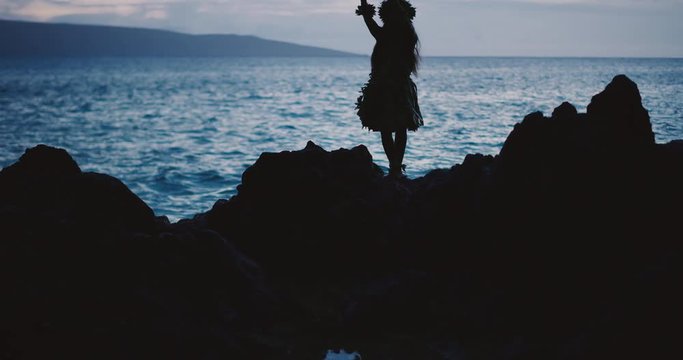 Silhouette of a traditional Hawaiian hula dancer woman dancing on a rugged island landscape at dusk in slow motion with an ocean background