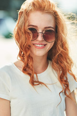 A beautiful red-haired girl in a white T-shirt and sunglasses walks down the street and smiles in...
