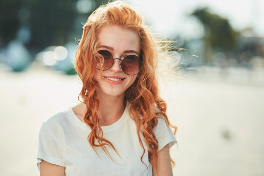 A beautiful red-haired girl in a white T-shirt and sunglasses walks down the street and smiles in frame