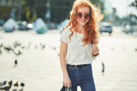A beautiful red-haired girl in a white T-shirt and sunglasses walks down the street and smiles in frame